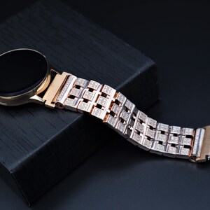 May include: Rose gold smartwatch with a black screen and a diamond-studded strap. The strap features a geometric pattern of square links, each encrusted with small, sparkling stones. The watch rests on a black textured surface.
