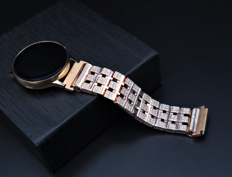 May include: Rose gold smartwatch with a black screen and a diamond-studded strap. The strap features a geometric pattern of square links, each encrusted with small, sparkling stones. The watch rests on a black textured surface.
