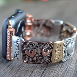 Puede incluir: Una correa de Apple Watch con un dise&ntilde;o decorativo. La correa presenta una mezcla de tonos plateados, cobrizos y dorados. Las palabras "Faith" y "Hope" son visibles, junto con acentos en forma de coraz&oacute;n. La correa est&aacute; sujeta a un Apple Watch negro.