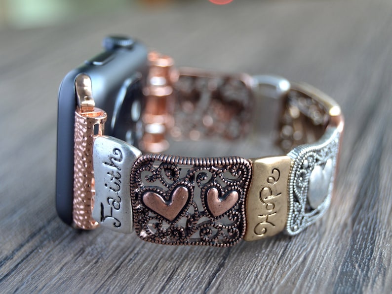 Puede incluir: Una correa de Apple Watch con un dise&ntilde;o decorativo. La correa presenta una mezcla de tonos plateados, cobrizos y dorados. Las palabras "Faith" y "Hope" son visibles, junto con acentos en forma de coraz&oacute;n. La correa est&aacute; sujeta a un Apple Watch negro.