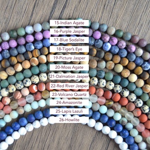 May include: A collection of 12 different types of gemstone beads, arranged in a semi-circle. The beads are all round and smooth, and they are arranged in rows of different colors. The beads are labeled with their names, such as Indian Agate, Purple Jasper, Blue Sodalite, Tiger's Eye, Picture Jasper, Moss Agate, Dalmation Jasper, Red River Jasper, Volcano Quartz, Amazonite, Lapis Lazuli, and Howlite.