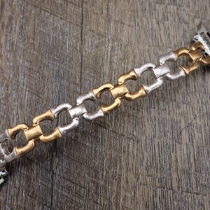 May include: A two-tone watch band with a chain-link design. The band alternates between gold and silver-colored links. The watch band is shown on a wooden surface.