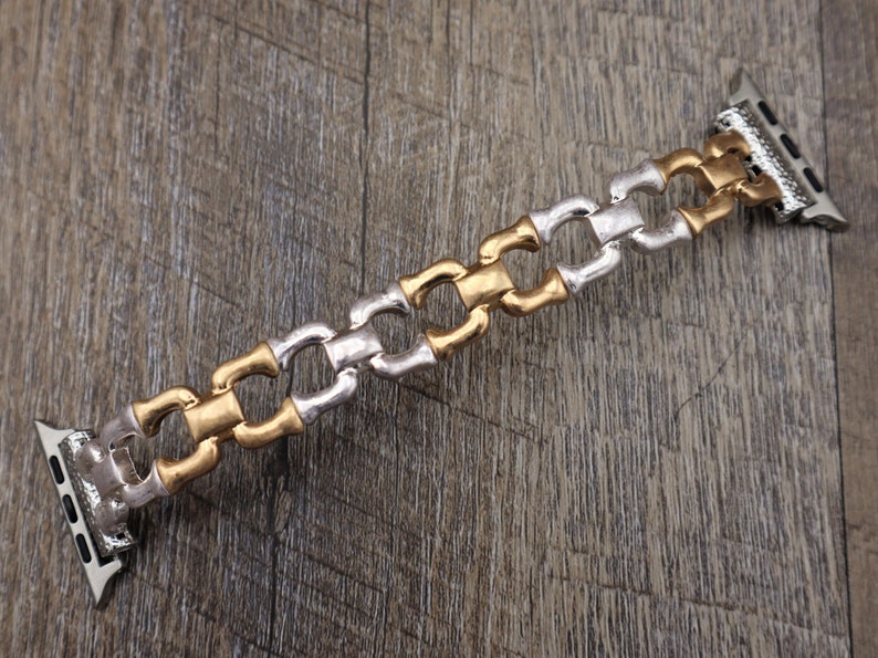 May include: A two-tone watch band with a chain-link design. The band alternates between gold and silver-colored links. The watch band is shown on a wooden surface.