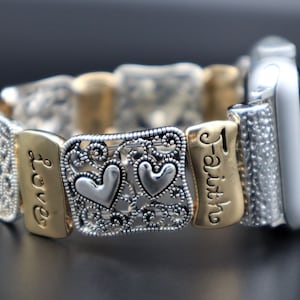 Puede incluir: Una banda de reloj Apple plateada y dorada con un delicado diseño de filigrana y dos dijes en forma de corazón. La banda tiene las palabras "Love" y "Faith" grabadas.