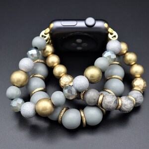 May include: A gray and gold beaded watch band with a black smartwatch. The band is made of multiple strands of beads, including gray, gold, and clear beads.