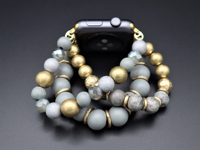 May include: A gray and gold beaded watch band with a black smartwatch. The band is made of multiple strands of beads, including gray, gold, and clear beads.