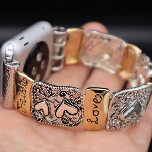 May include: A silver and gold Apple Watch band with a decorative design featuring hearts and the words "Faith" and "Love".