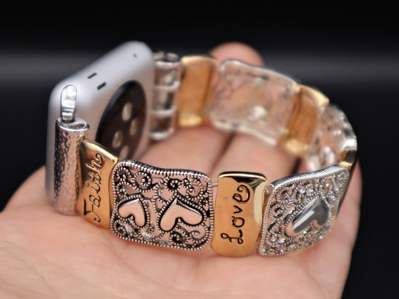 May include: A silver and gold Apple Watch band with a decorative design featuring hearts and the words "Faith" and "Love".