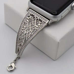 May include: A silver watch band with a decorative, intricate design featuring a repeating pattern of swirls and diamonds. The band has a clasp closure.
