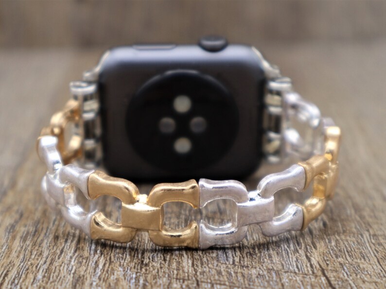 May include: A two-tone watch band with a chain link design. The links alternate between gold and silver colors. The watch band is attached to a black smartwatch.