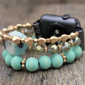 May include: A black Apple Watch with a gold and turquoise beaded bracelet. The bracelet has three layers of beads, including gold beads, clear crystal beads, and turquoise wooden beads.