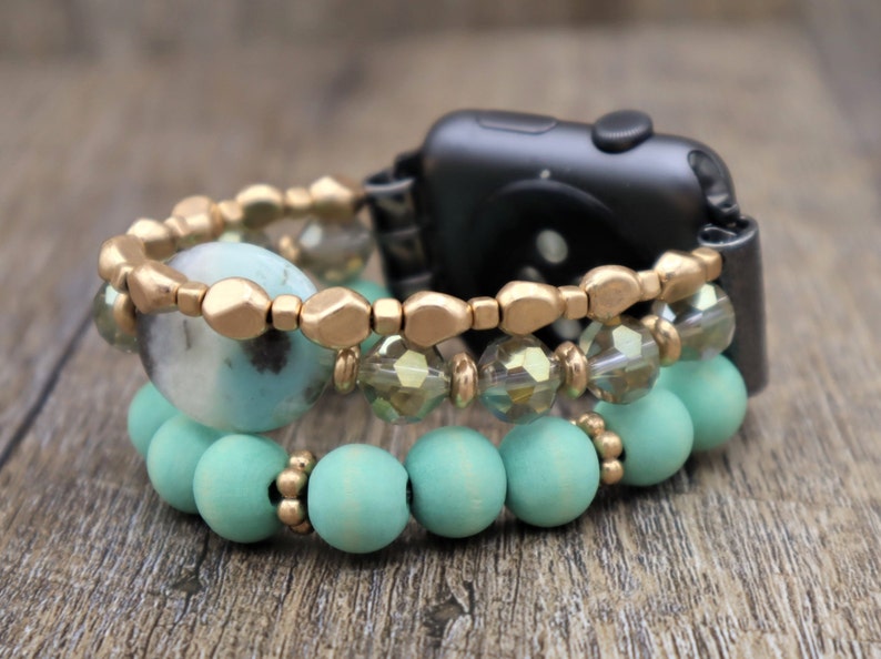 May include: A black Apple Watch with a gold and turquoise beaded bracelet. The bracelet has three layers of beads, including gold beads, clear crystal beads, and turquoise wooden beads.