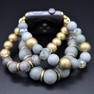 Puede incluir: Una pulsera de reloj de cuentas grises, doradas y transparentes con un cierre dorado. La pulsera está hecha de cuentas de varios tamaños y formas, incluyendo redondas, ovaladas e irregulares.