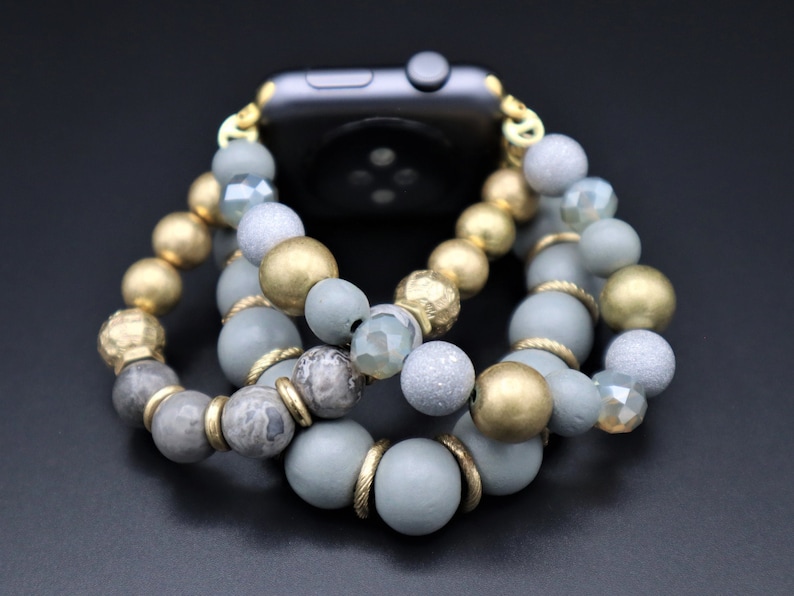 May include: A gray, gold, and clear beaded watch band with a gold clasp. The band is made of various sizes and shapes of beads, including round, oval, and irregular shapes.