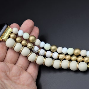 K&ouml;nnte beinhalten: Ein goldfarbenes und wei&szlig;es Armband aus Perlen f&uuml;r die Apple Watch mit einem goldfarbenen Verschluss. Das Armband besteht aus drei Reihen Perlen, wobei die obere Reihe wei&szlig;, die mittlere Reihe goldfarben und die untere Reihe wei&szlig; ist.