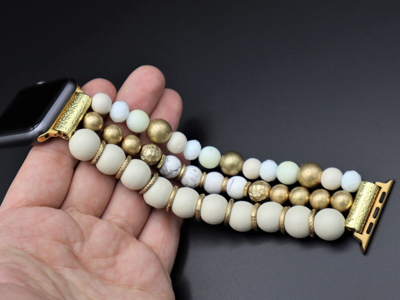 K&ouml;nnte beinhalten: Ein goldfarbenes und wei&szlig;es Armband aus Perlen f&uuml;r die Apple Watch mit einem goldfarbenen Verschluss. Das Armband besteht aus drei Reihen Perlen, wobei die obere Reihe wei&szlig;, die mittlere Reihe goldfarben und die untere Reihe wei&szlig; ist.