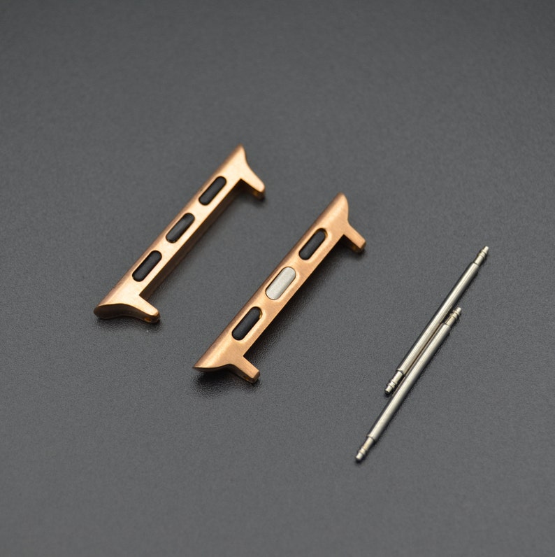May include: Two rose gold watch band adapters with black and white accents, and two silver spring bars. The adapters have a rectangular shape with rounded edges and are designed to connect a watch band to a watch case.