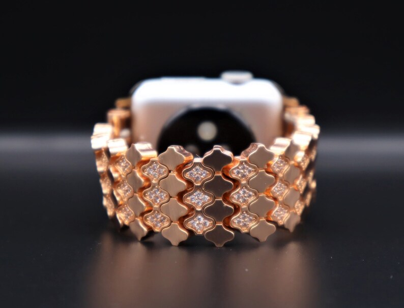 May include: A rose gold Apple Watch band with a diamond-like pattern. The band is made of metal links and has a clasp closure.