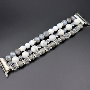 May include: A silver and grey beaded Apple Watch band with three rows of beads. The beads are white, grey, and clear.