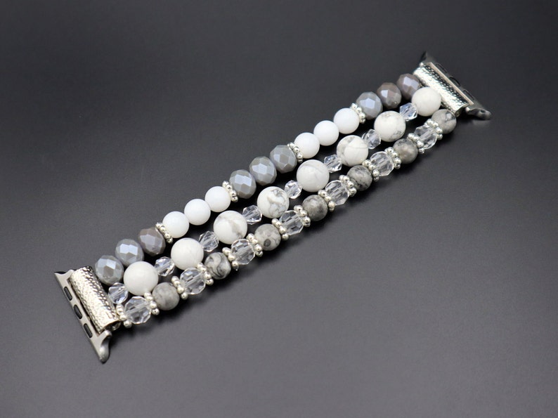 May include: A silver and grey beaded Apple Watch band with three rows of beads. The beads are white, grey, and clear.