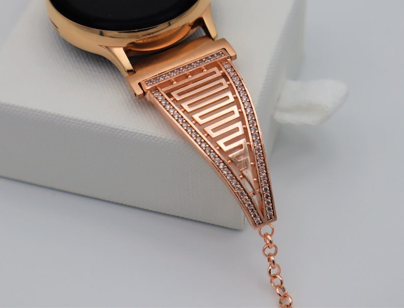 May include: Rose gold watch strap with a geometric design and clear rhinestones. The strap is attached to a rose gold watch.