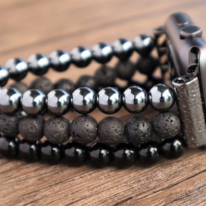 Puede incluir: Un reloj inteligente negro con una banda de cuentas. La banda está hecha de tres capas de cuentas: negras, grises y negras. Las cuentas son redondas y tienen un acabado mate.