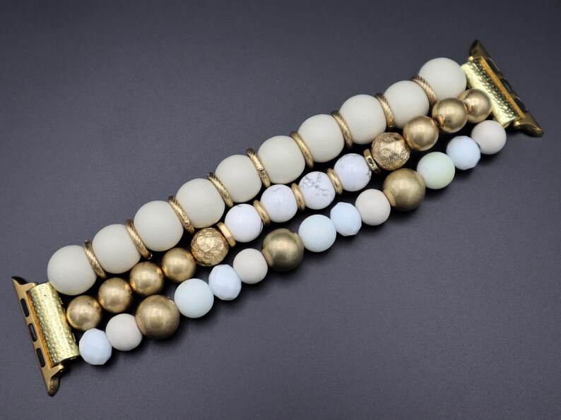 K&ouml;nnte beinhalten: Ein goldfarbenes und wei&szlig;es Armband f&uuml;r die Apple Watch mit dreireihigem Design. Das Armband besteht aus wei&szlig;en, cremefarbenen und hellgr&uuml;nen Perlen sowie goldfarbenen Metallperlen.