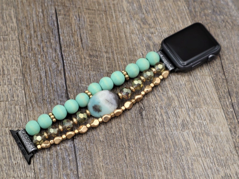 May include: A turquoise and gold beaded Apple Watch band with a large, oval-shaped gemstone. The band is made of wooden beads, gold beads, and clear crystal beads.