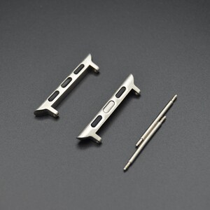 May include: Two silver-toned watch band adapters with black accents and two silver spring bars. The adapters have a sleek, modern design, and are likely for a smartwatch. The items are displayed on a dark gray surface.