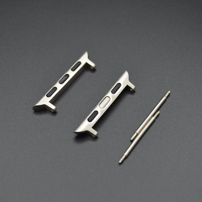 May include: Two silver-toned watch band adapters with black accents and two silver spring bars. The adapters have a sleek, modern design, and are likely for a smartwatch. The items are displayed on a dark gray surface.