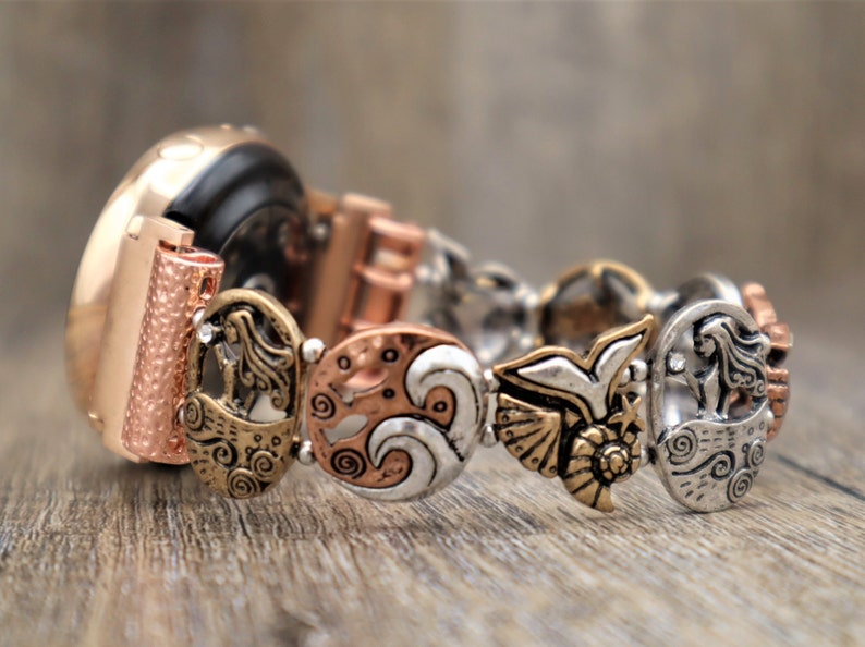 May include: A rose gold smartwatch with a decorative metal band featuring a mermaid, waves, and a bird design. The band is made of mixed metals with silver, gold, and copper tones.