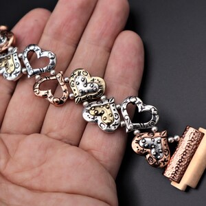 May include: A silver, gold, and copper bracelet with a series of open heart charms. The bracelet has a clasp closure.