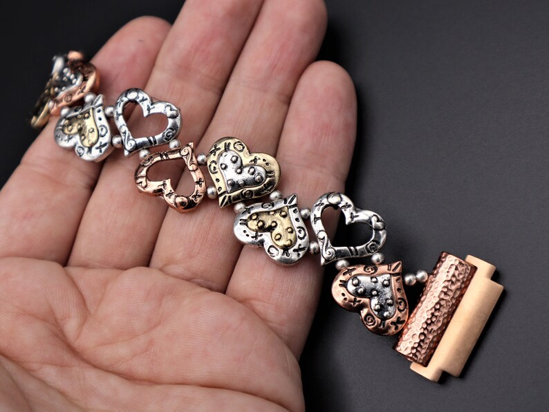 May include: A silver, gold, and copper bracelet with a series of open heart charms. The bracelet has a clasp closure.