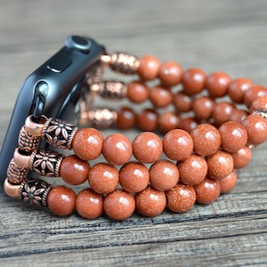 Puede incluir: Una pulsera de reloj de cuentas de piedra arenisca marrón con detalles en cobre. La pulsera está envuelta alrededor de un reloj inteligente negro.