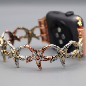 Puede incluir: Una banda de Apple Watch de oro rosa con un diseño decorativo de estrella de mar. La banda está hecha de metal y tiene una mezcla de tonos plateados, dorados y cobrizos.