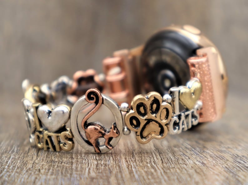 May include: A silver and gold charm bracelet with cat-themed charms, including a paw print, a heart with "I Love Cats" text, and a cat silhouette. The bracelet is attached to a rose gold smartwatch.