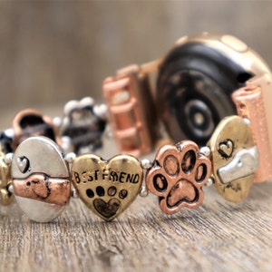 Puede incluir: Una pulsera el&aacute;stica con varios dijes de metal. Los dijes incluyen un coraz&oacute;n con el texto "BEST FRIEND", una huella de pata y dise&ntilde;os con tem&aacute;tica de perros. La pulsera tiene tonos dorados, plateados y cobrizos y est&aacute; sobre una superficie de madera.