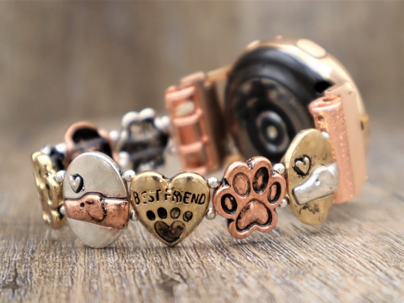 Puede incluir: Una pulsera el&aacute;stica con varios dijes de metal. Los dijes incluyen un coraz&oacute;n con el texto "BEST FRIEND", una huella de pata y dise&ntilde;os con tem&aacute;tica de perros. La pulsera tiene tonos dorados, plateados y cobrizos y est&aacute; sobre una superficie de madera.