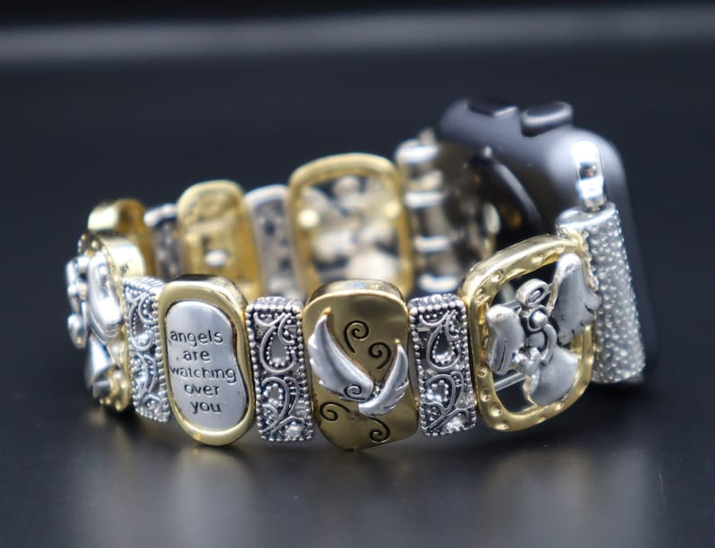 May include: Gold and silver tone watch band with a charm bracelet style. The charms feature angel wings, an angel, and the text "angels are watching over you".