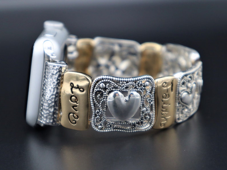 May include: Silver and gold Apple Watch band with a heart charm and engraved words "Love" and "Faith".
