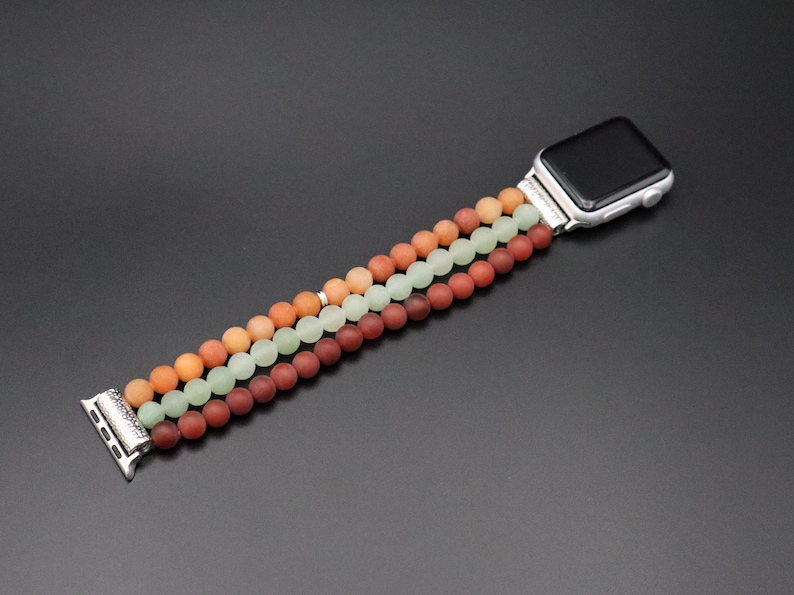 May include: A three-strand beaded Apple Watch band with red, green, and orange beads. The band is made of smooth, round beads and has a silver clasp.