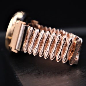 K&ouml;nnte beinhalten: Eine goldfarbene Uhr mit einem runden Zifferblatt und einem einzigartigen, kunstvoll gestalteten Armband. Das Armband besteht aus miteinander verbundenen, &uuml;berlappenden, gebogenen Teilen, die ein wellenf&ouml;rmiges Muster erzeugen.