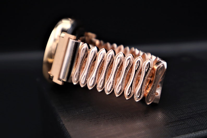 K&ouml;nnte beinhalten: Eine goldfarbene Uhr mit einem runden Zifferblatt und einem einzigartigen, kunstvoll gestalteten Armband. Das Armband besteht aus miteinander verbundenen, &uuml;berlappenden, gebogenen Teilen, die ein wellenf&ouml;rmiges Muster erzeugen.
