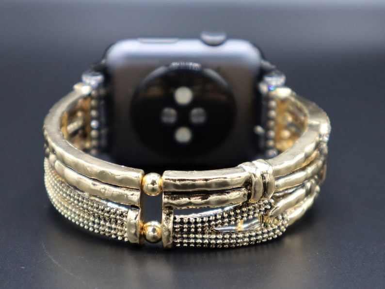 May include: Gold-toned watch band with a textured, hammered metal design. The band has a decorative, intricate pattern with small beads and a clasp closure.