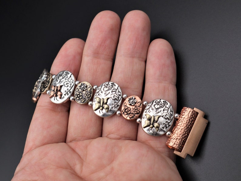 Pode incluir: Um bracelete de prata e cobre com um design floral e detalhes de borboleta. O bracelete tem um fecho de encaixe.