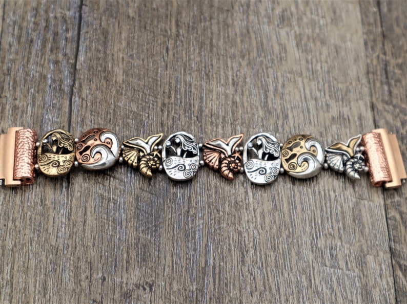 May include: A silver and copper bracelet with a mermaid and wave design. The bracelet is made of metal and has a clasp closure.