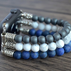 Puede incluir: Un reloj inteligente negro con una banda de cuentas plateadas. La banda está hecha de tres filas de cuentas: gris oscuro, blanco y azul.