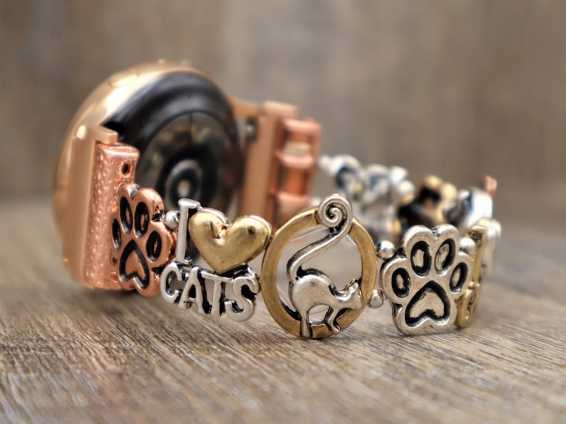 May include: A rose gold smartwatch with a charm bracelet attachment. The charms include paw prints, a heart with "I love cats" written on it, a cat, and more paw prints.