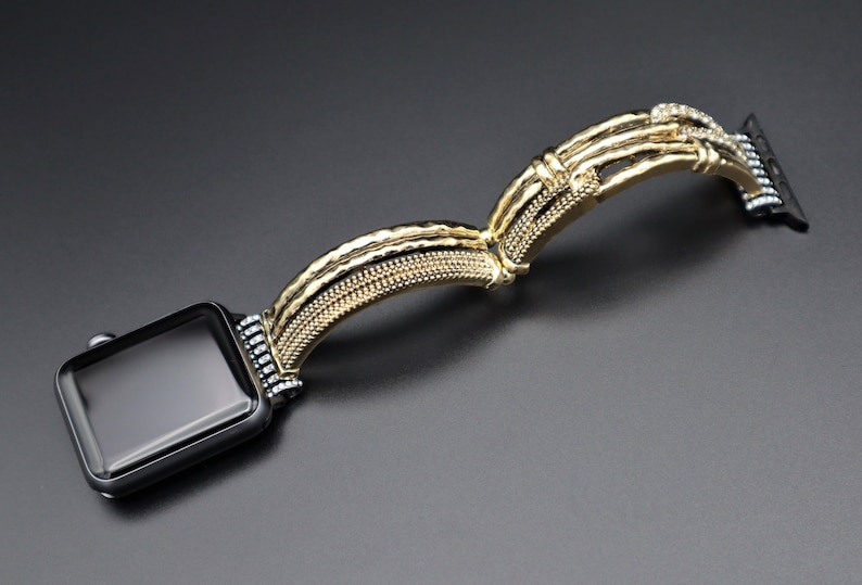 May include: A gold-toned watch band with a textured design and clear rhinestones. The band is designed to fit a smart watch.