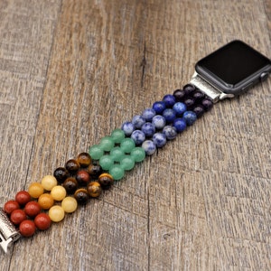 Op de afbeelding: Een kleurrijk kralenbandje voor de Apple Watch met rode, oranje, gele, groene, blauwe en paarse kralen. Het bandje is gemaakt van natuurlijke stenen en is ontworpen om te passen op een standaard Apple Watch.